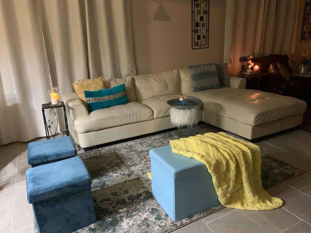 Cozy living room with a beige sectional sofa, blue ottomans, a yellow throw, candles, and a patterned rug