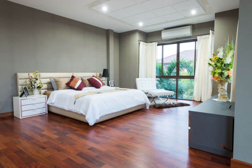 Spacious gray bedroom with hardwood floors, large bed, and white ottoman by window.