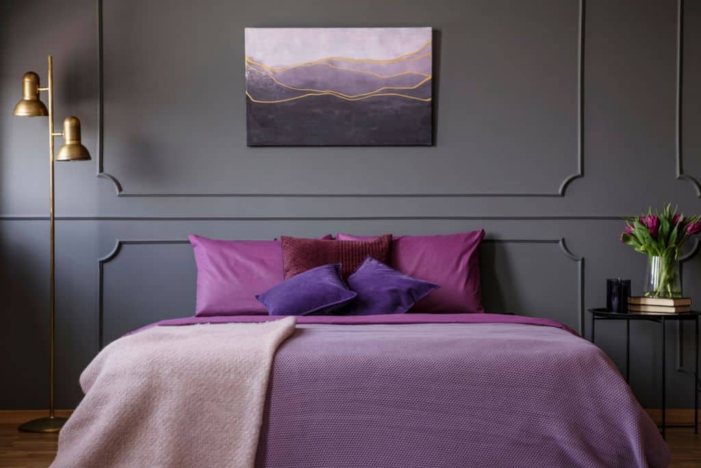 Modern bedroom with purple bedding, dark gray walls, and gold floor lamp.