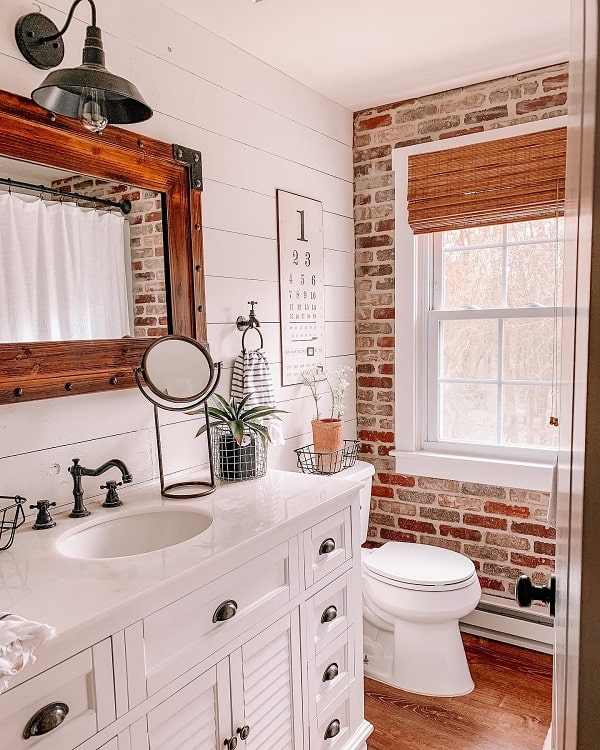 A farmhouse-style bathroom with a wooden mirror, potted plants, and a brick accent wall near a window with bamboo shades