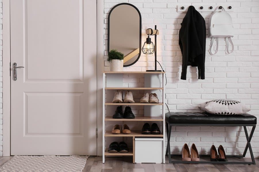 Entryway setup with a stylish wooden shoe rack, black bench, wall hooks, and a decorative mirror on a white brick wall