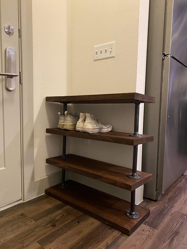 Industrial-style wooden shoe rack with three shelves and metal pipe supports, placed in a corner near a door on hardwood flooring