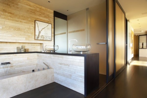 Modern bathroom with large built-in tub, glass vessel sinks, and beige stone walls.