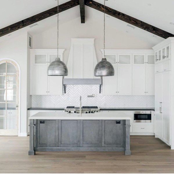 Charming kitchen with rustic silver metal pendant lights hanging above a gray island, complemented by white cabinetry, a herringbone backsplash, and exposed wooden beams