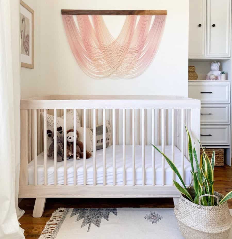 Modern baby room with light wood crib, pink hanging wall decor, and potted snake plant.