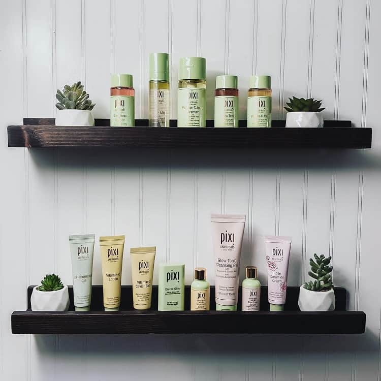 Black bathroom shelves displaying Pixi beauty products and small succulents against a white wall.