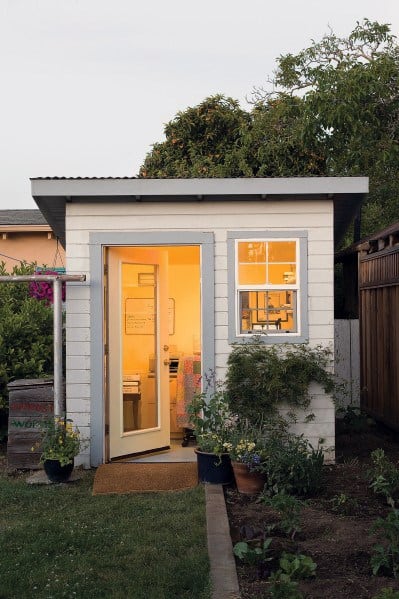 Small office shed with glass door, white exterior, and window, lit from inside.