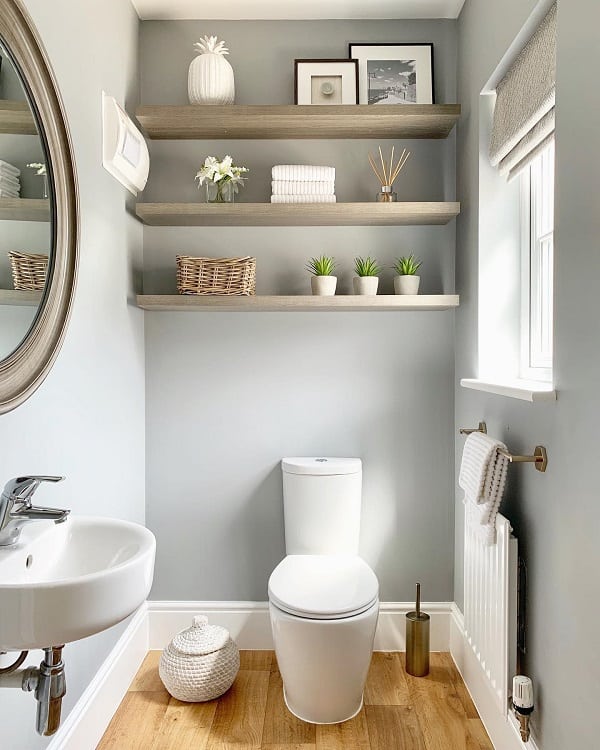 Minimalist gray bathroom with floating wooden shelves, round mirror, and small potted plants.