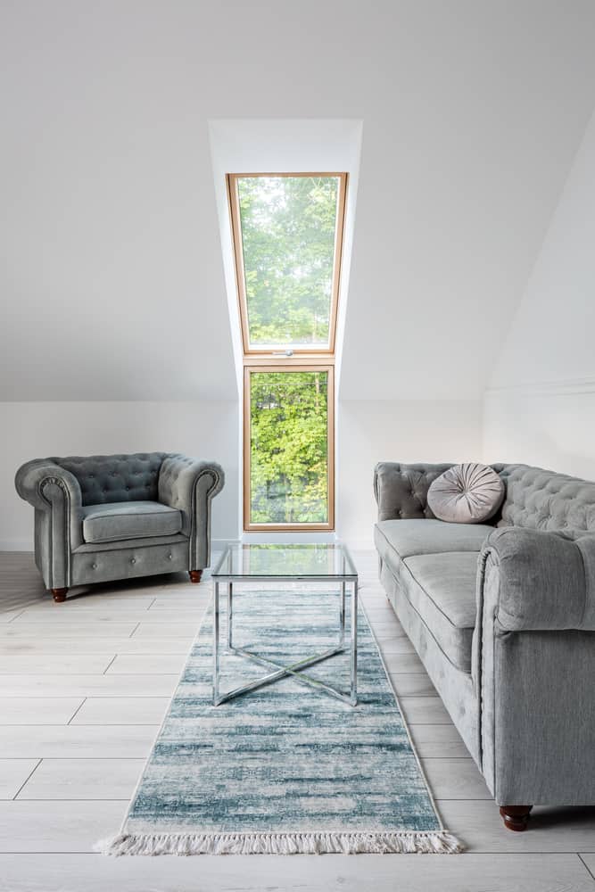 Chic attic living room with a skylight, tufted armchairs, glass coffee table, and a soft blue rug for a serene vibe