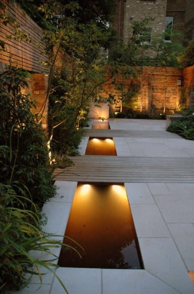 Narrow backyard side pond with soft lighting, surrounded by modern patio tiles, greenery, and brick walls.