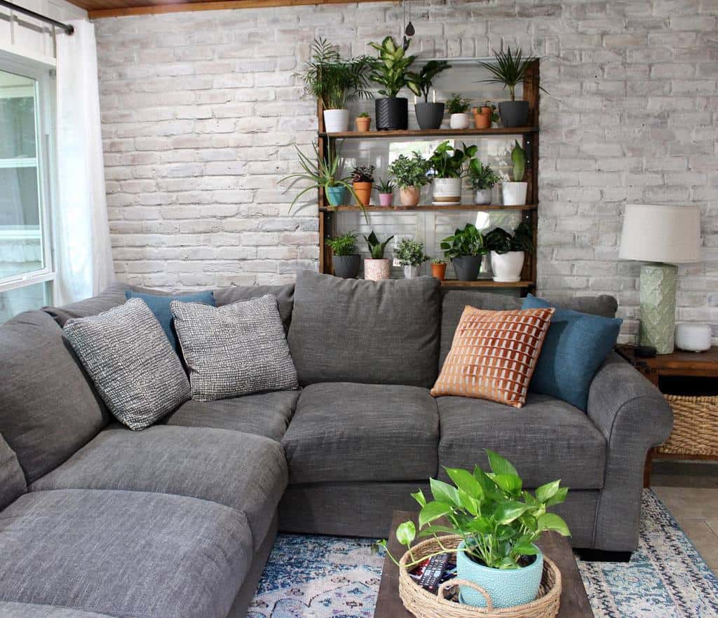 Cozy sunroom with gray sectional sofa, potted plants, white brick wall, and wooden shelves.