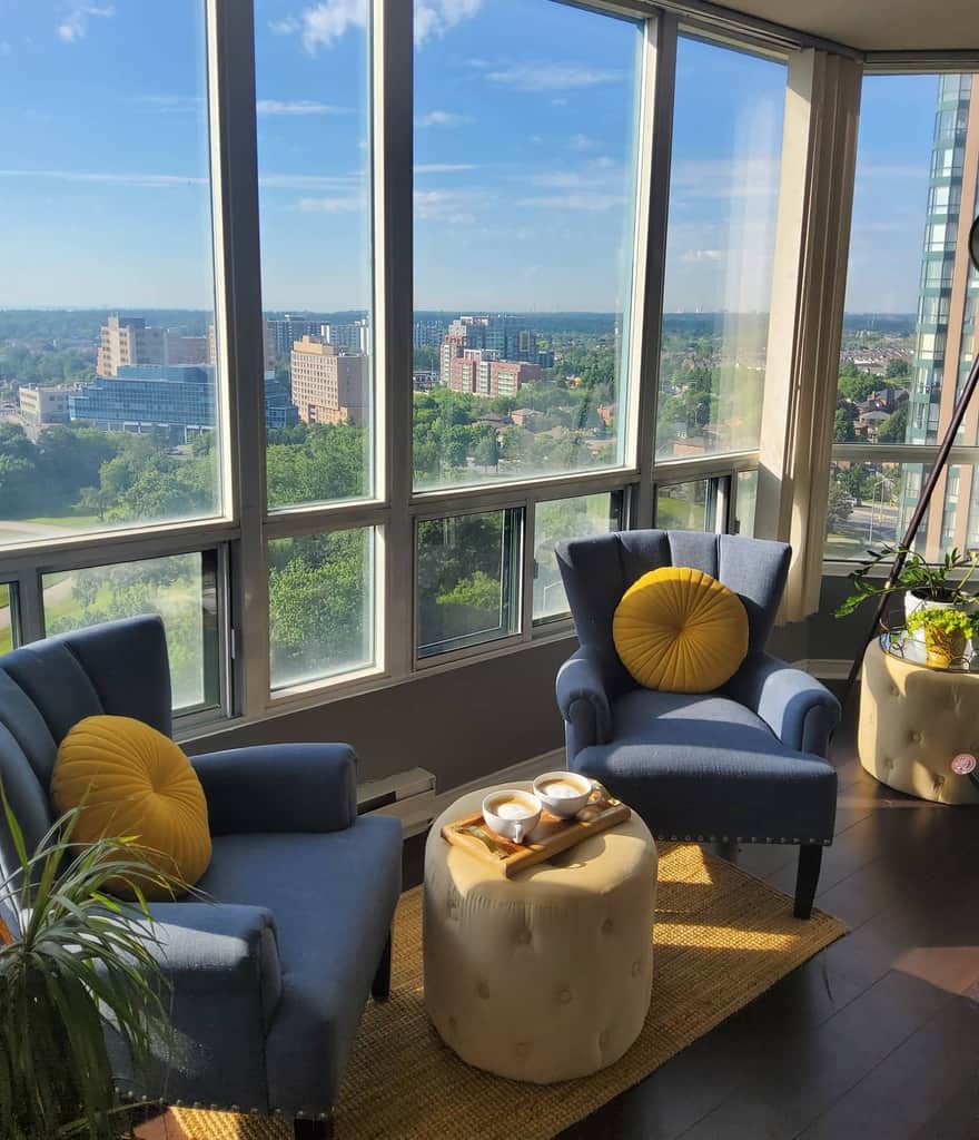 Cozy sunroom with blue armchairs, yellow pillows, round ottomans, and a warm, inviting atmosphere.