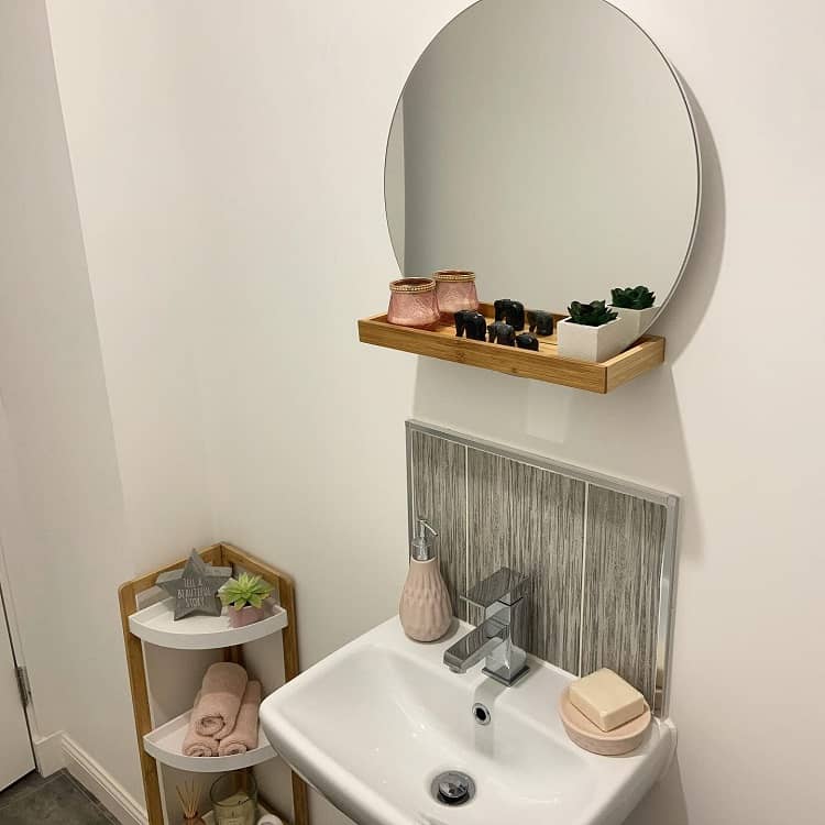 Small bathroom sink with round mirror, wooden shelf, and corner storage unit for towels and decor.