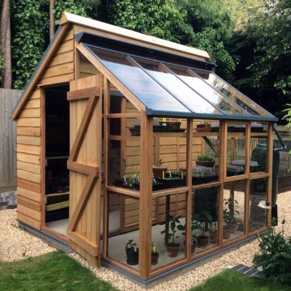 Wooden greenhouse shed with glass walls and roof, designed for gardening in a landscaped yard.