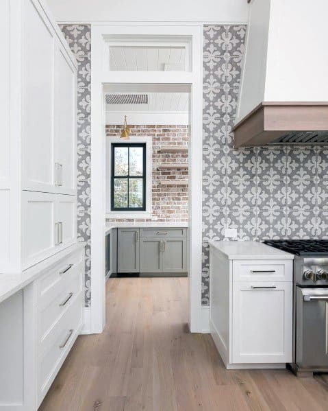 Sleek kitchen with light wood flooring, white cabinetry, and gray patterned tile wall featuring exposed brick