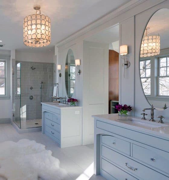 Elegant bathroom with light blue vanities, oval mirrors, glass shower, and a chic chandelier.