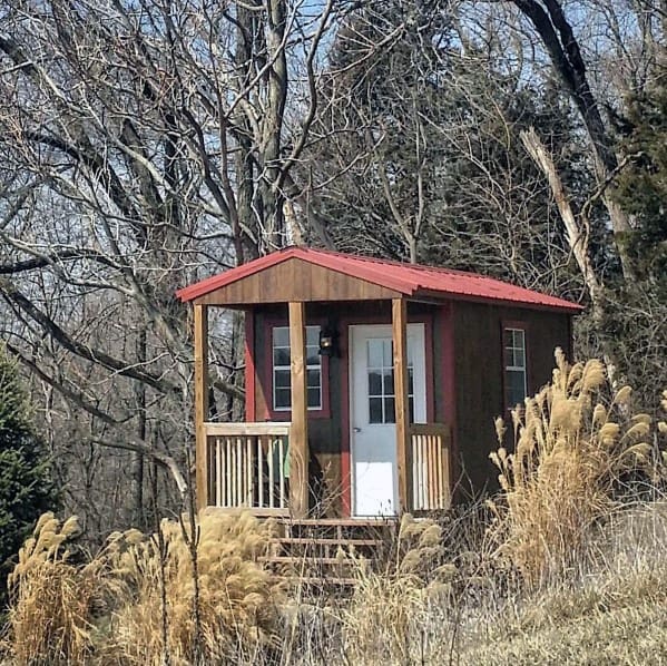 Small wooden shed with a red metal roof and porch, surrounded by tall grasses.