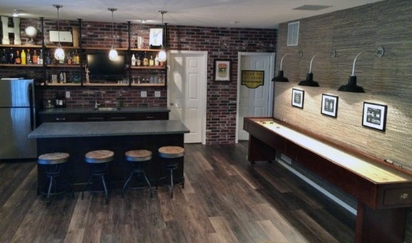 Rustic man cave with brick walls, industrial-style wet bar, bar stools, and shuffleboard table.