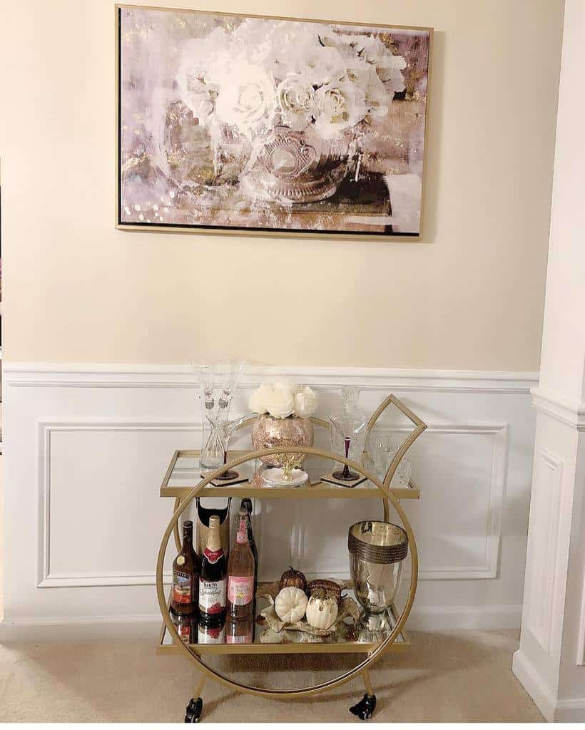 Small gold bar cart with champagne bottles, pumpkins, and elegant glassware under floral artwork.