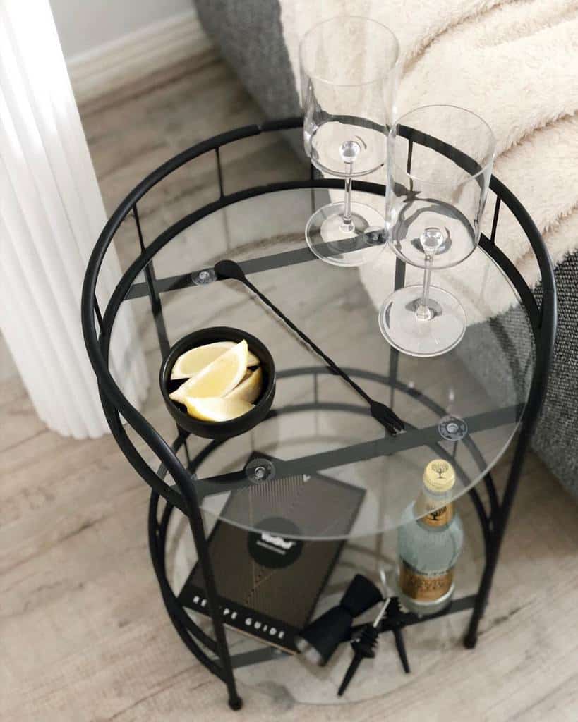 Small black metal and glass bar cart with wine glasses, lemon slices, and a bottle of tonic water.