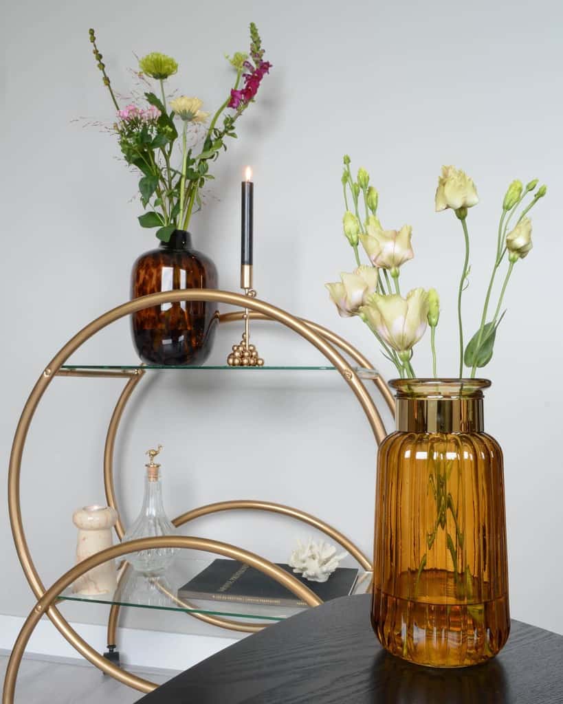 Small gold bar cart with amber vase, flowers, candle, and decorative items on glass shelves.