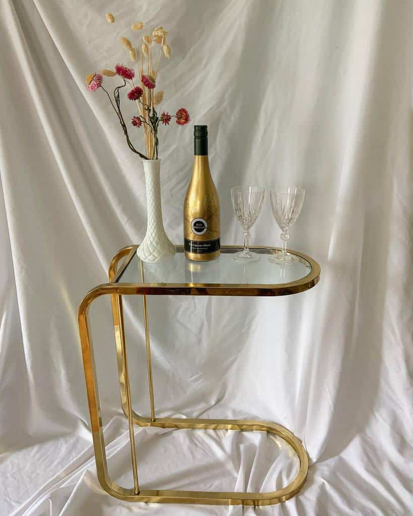 Small gold bar cart with wine bottle, glasses, and vase of dried flowers against white backdrop.