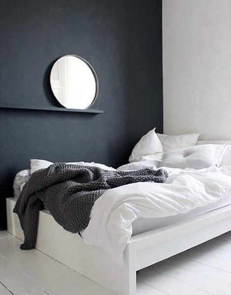 Modern bedroom with black accent wall, round mirror, and cozy white bedding with gray throw.