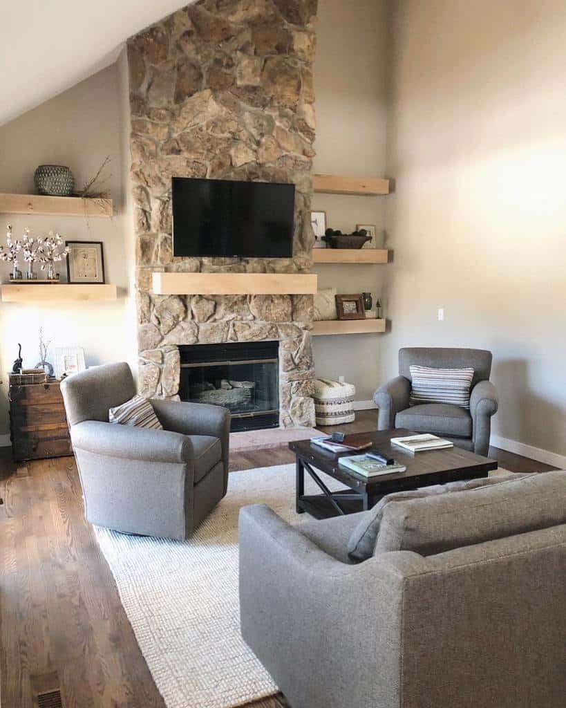 Cozy living room with a stone fireplace, TV, gray armchairs, and wooden shelves adorned with decor