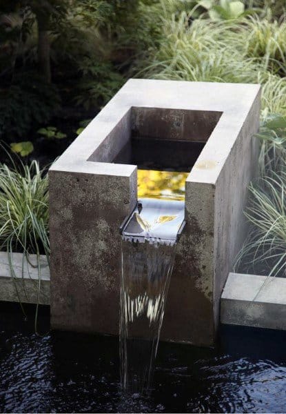 Concrete waterfall feature with a minimalist design, cascading into a small pond surrounded by plants.