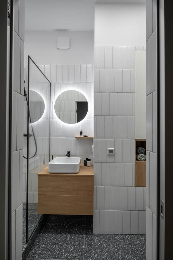 Contemporary bathroom with backlit round mirrors, wood floating vanity, and white tile walls.