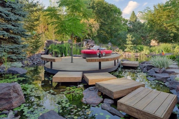 Ecosystem pond with floating wooden platforms, lily pads, and a circular deck featuring seating, surrounded by lush greenery.