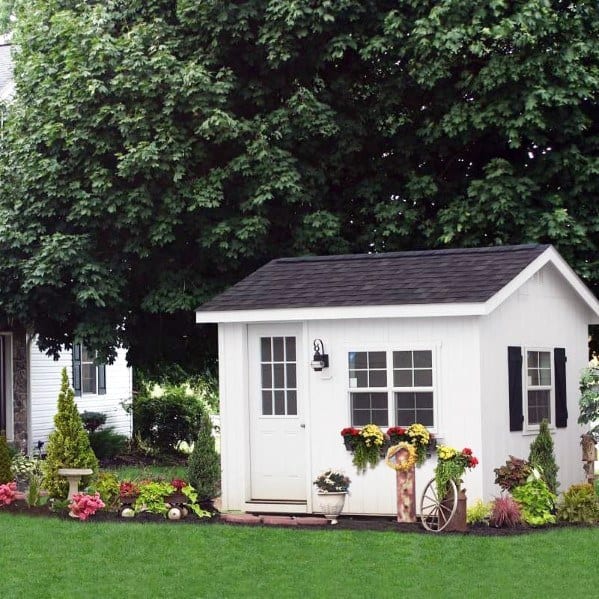 White garden shed with black shutters, flower pots, and decor in a landscaped backyard.