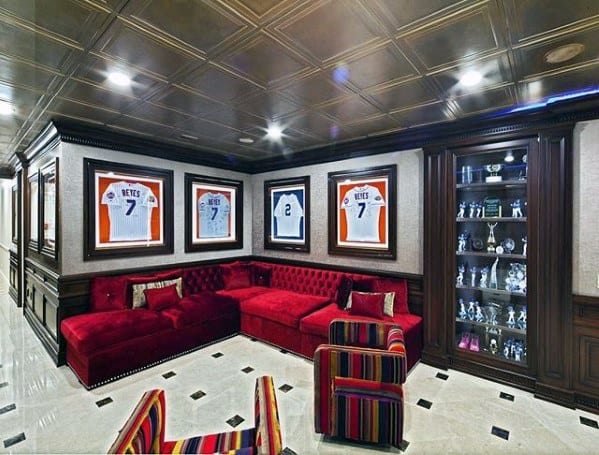 Luxurious sports-themed mancave with red velvet sectional, framed jerseys, striped chairs, and trophy display.