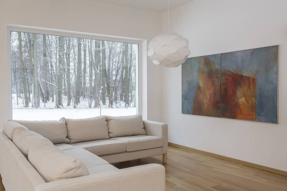 Bright minimalist living room with cream sectional sofa, artistic pendant light, abstract wall art, and large window overlooking a snowy forest