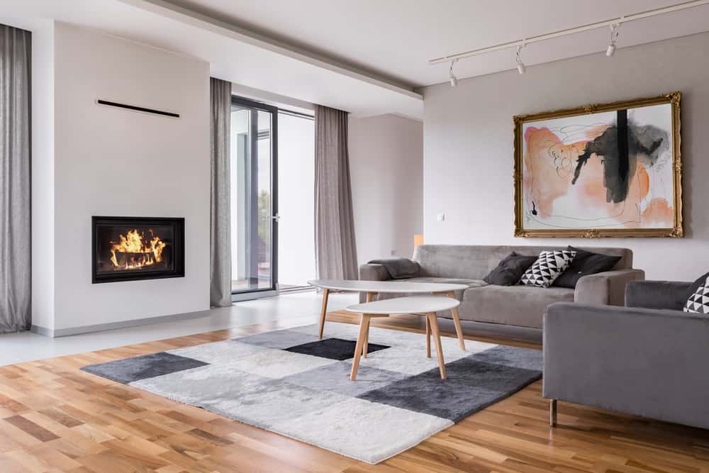 Minimalist living room with a gray sofa, modern fireplace, abstract framed artwork, geometric rug, and large windows with sheer curtains