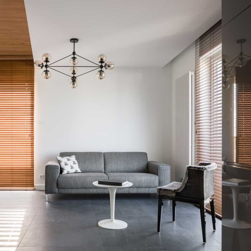 Minimalist living room featuring a gray sofa, modern white side table, unique chandelier, and wooden blinds for natural lighting