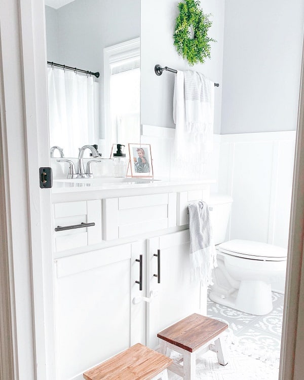 Farmhouse-style bright white bathroom with a towel, green wreath, and wooden stools in front of the sink