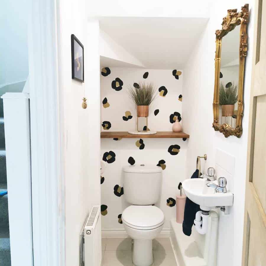 Small, stylish bathroom with a toilet, sink, and an accent wall; gold-framed mirror and shelf with plants