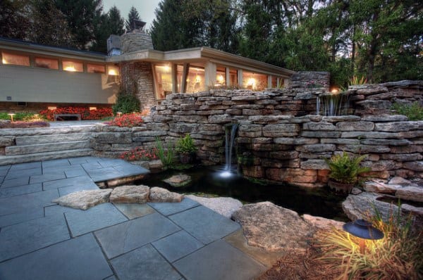 Natural tiered pond with stone waterfall feature, illuminated at night, surrounded by a modern patio and landscaping.