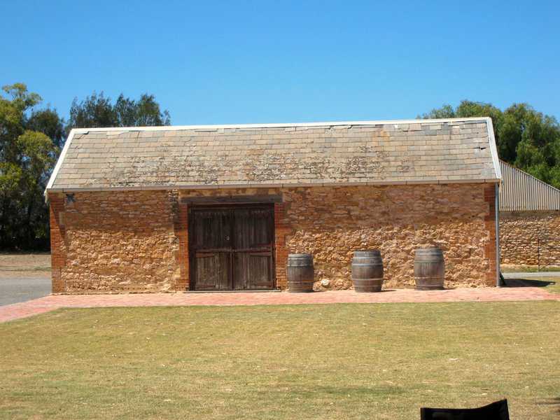 Rustic stone cottage shed with a weathered roof, wooden doors, and barrels on a brick patio.