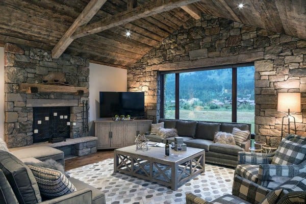 A rustic living room with a stone fireplace, exposed wooden beams, cozy seating, and large windows offering scenic views, creating a warm and inviting atmosphere