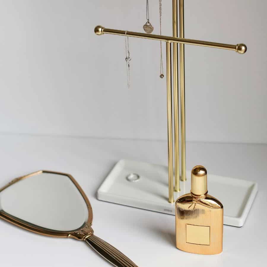 Gold jewelry stand with mirror and perfume bottle on a white surface.