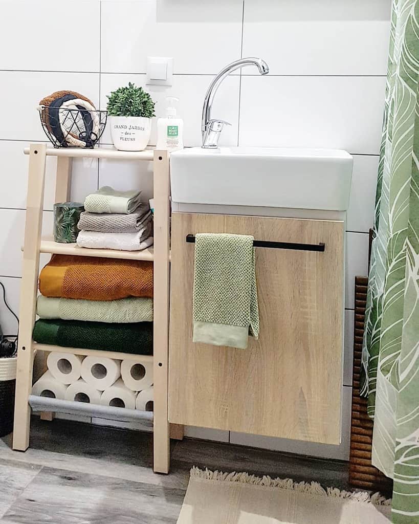 Small bathroom sink with wooden storage rack, folded towels, toilet paper, and decorative items.