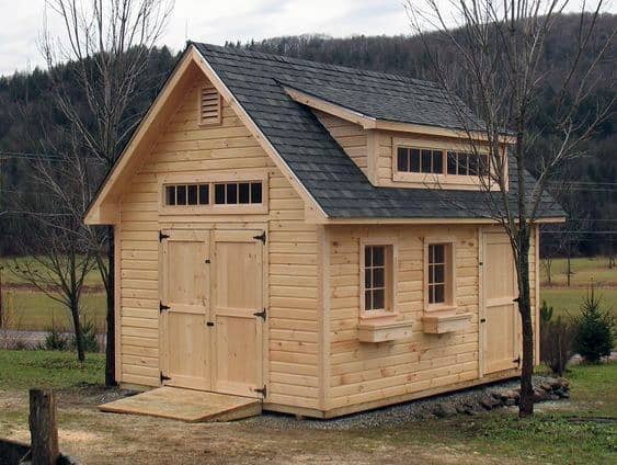 Natural wood shed with farmhouse-style doors, a shingled roof, and window boxes in a rural landscape.