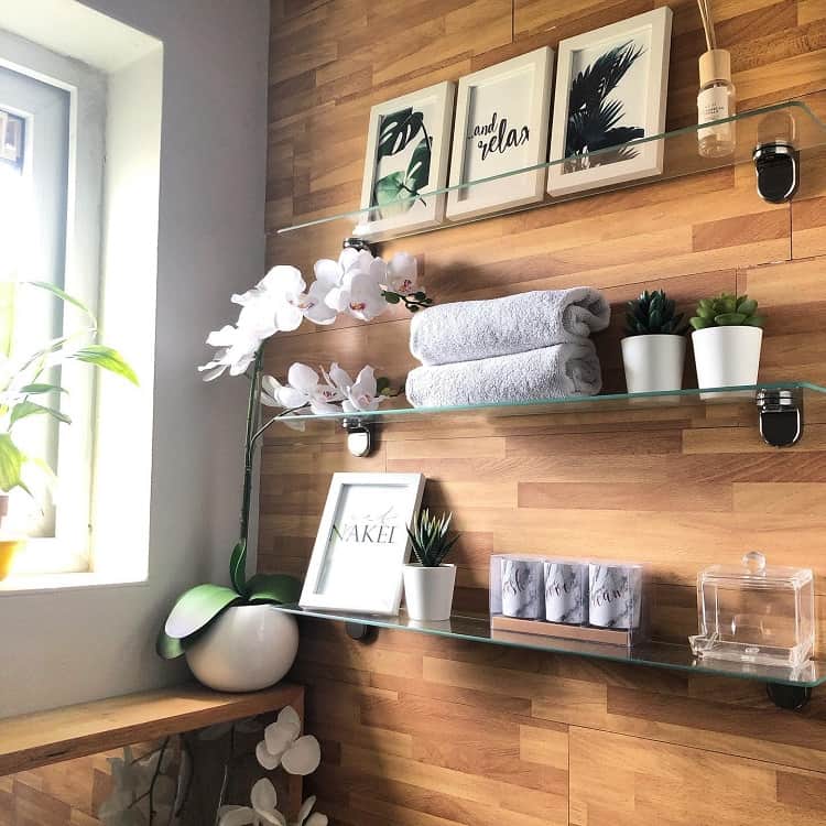 Glass bathroom shelves with folded towels, potted plants, and framed wall art against a wooden wall.