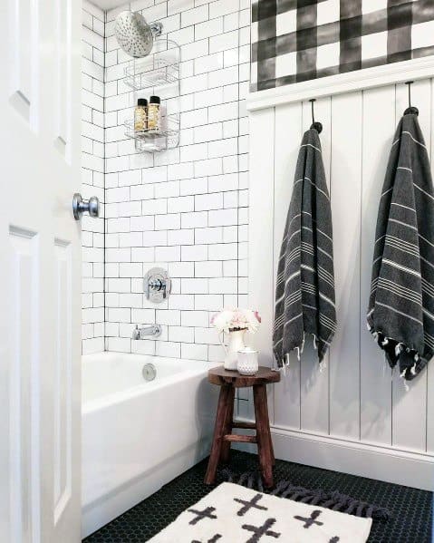 White bathroom with black and white towels, bathtub, stool with flowers, and cross-patterned bath mat