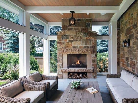 This sunroom features a rustic stone fireplace, natural wood ceiling, large glass windows, and cozy wicker seating with neutral cushions.