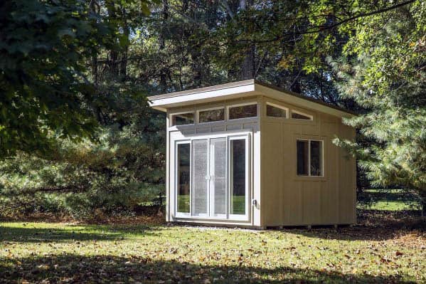 Modern shed with large glass doors and a slanted roof, surrounded by trees and foliage.