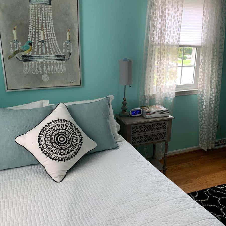 Turquoise bedroom with patterned pillow, chandelier artwork, and lace curtains.