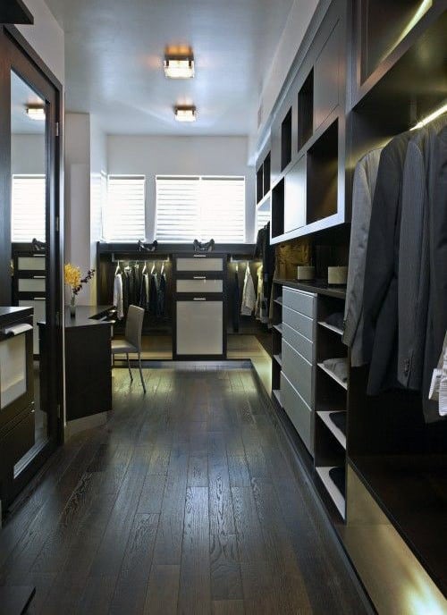 Modern walk-in closet with dark wood flooring, shelves, hanging clothes, and a chair by the window